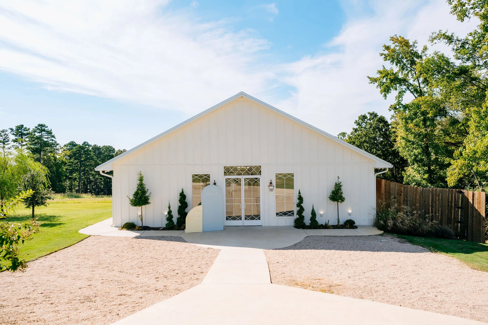 A white building with large windows and glass double doors is surrounded by small trees, with a gravel pathway leading up to it. There is a blue mailbox and a small sign in front of the building, and a wooden fence on the right side. The sky is partly cloudy and the trees are green.